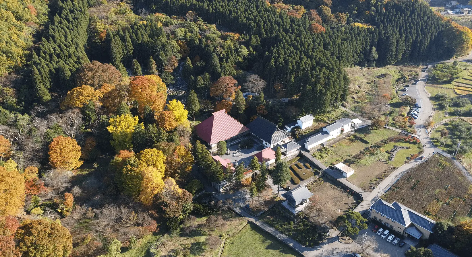 紅葉が広がる山あいにたたずむ岩松院の全景を上空から撮影。赤い本堂が秋の自然に映える
