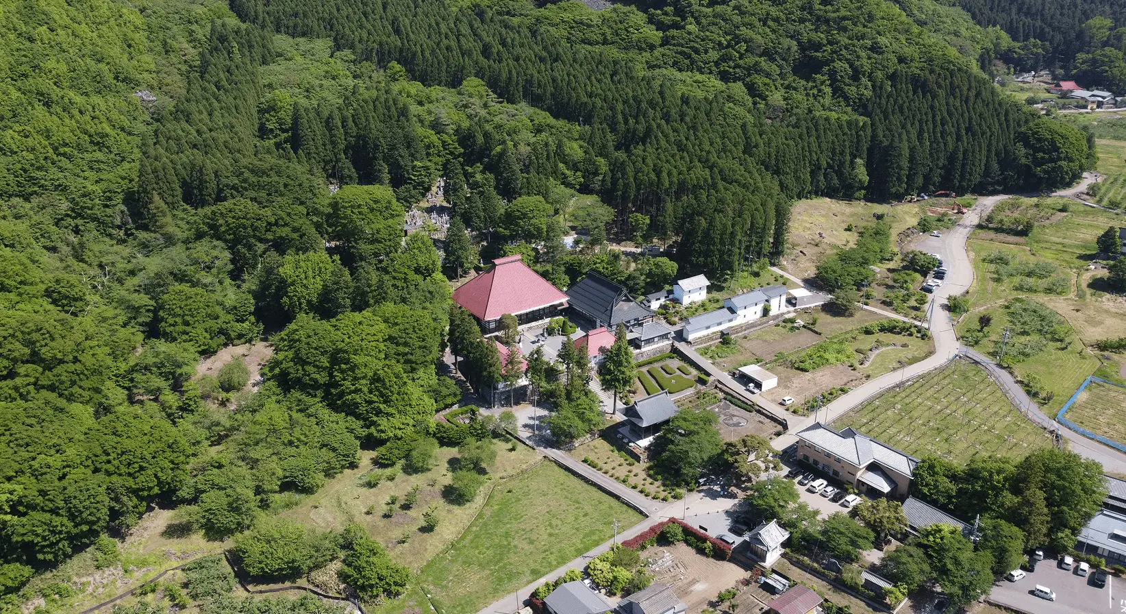 濃い緑に包まれた夏の岩松院全景を上空から望む。赤い本堂が森の中に映える