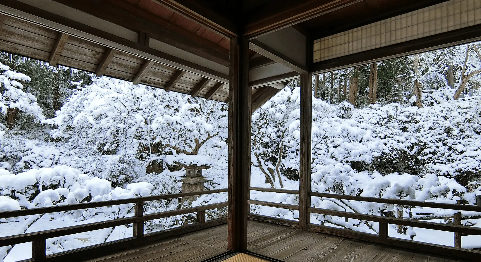 雪化粧した庭を縁側から眺める冬の岩松院。木造の柱越しに、雪の枝や石灯籠が見える静寂な風景