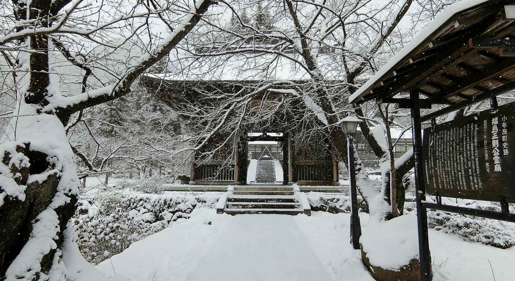 雪に覆われた参道と山門。木々の枝にも雪が積もり、歴史を感じさせる厳かな冬の岩松院の入口